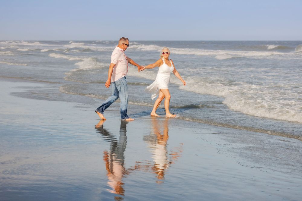 Elderly couple holding hands and walking barefoot along a sunny beach shoreline with ocean waves.