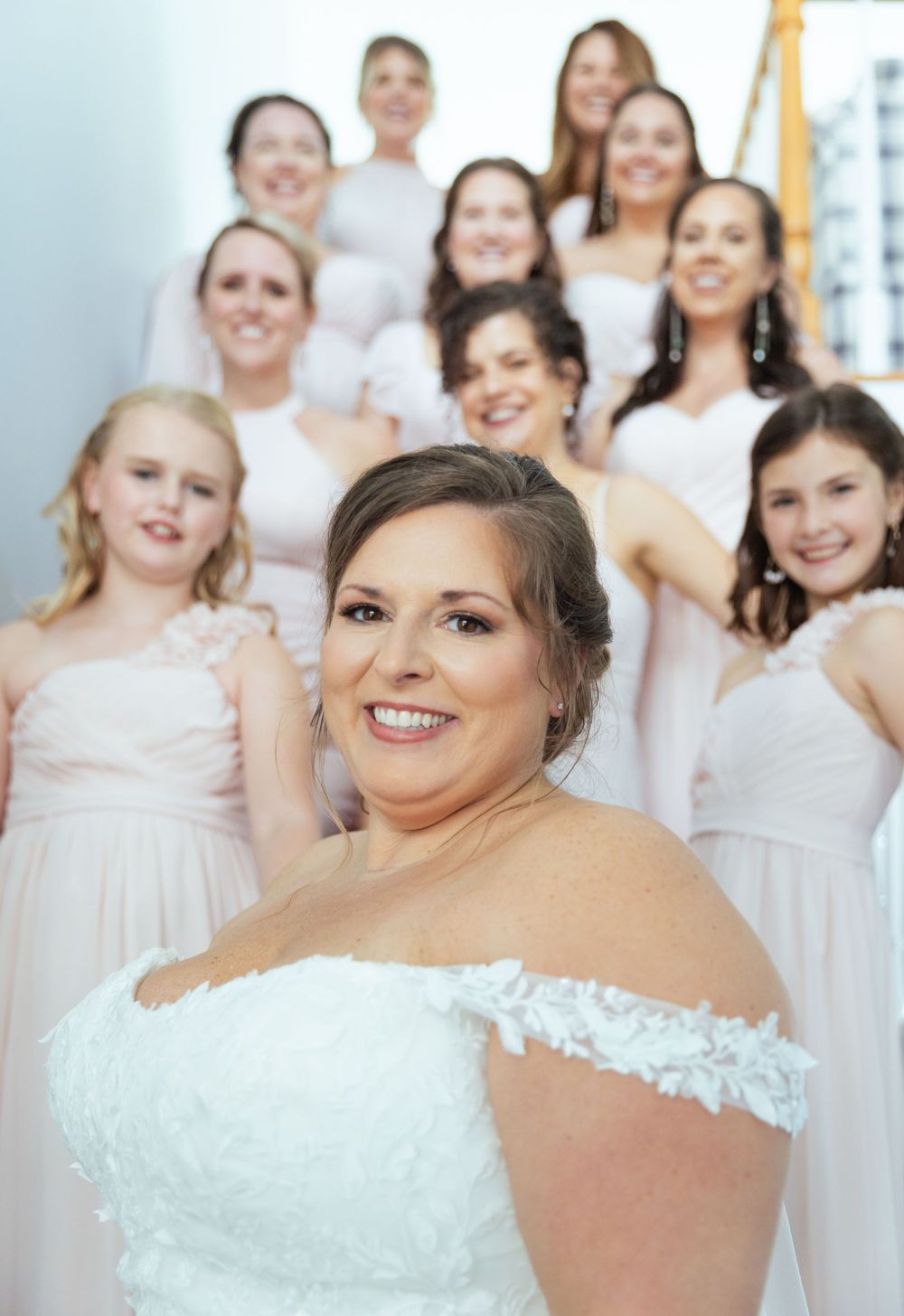 Bride in white lace gown smiling with bridesmaids in blush dresses on staircase.