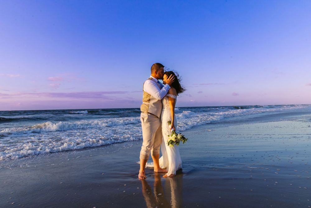 Newlywed couple embraces on a wet beach shore at twilight with ocean waves behind them.