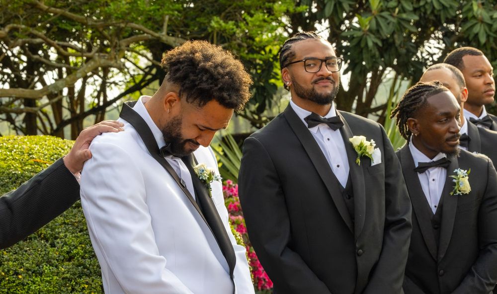 Groomsmen in tuxedos with boutonnieres share an emotional moment outdoors at a wedding ceremony.