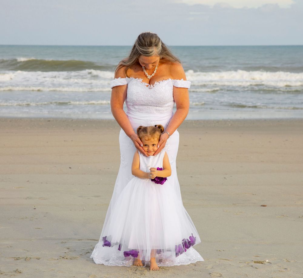 Bride in white off-shoulder gown with flower girl in white dress with purple accents on a sandy beach.