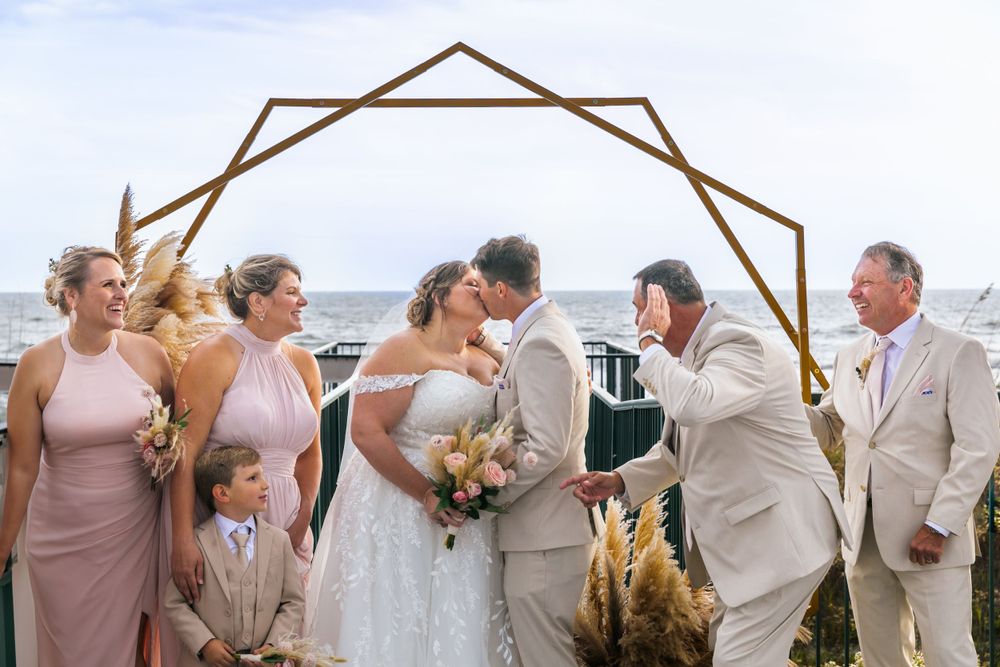 Wedding couple kissing under geometric arch with bridal party at oceanside ceremony.