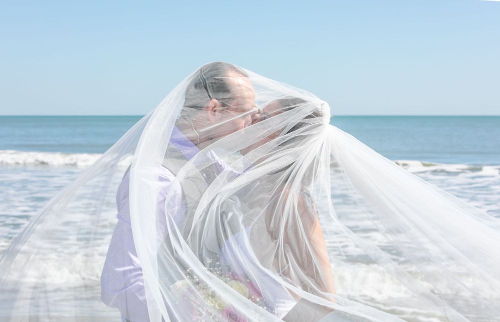 Bride wrapped in flowing white veil on beach with ocean waves in background on sunny day.