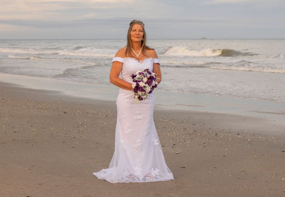 Bride in white off-shoulder mermaid gown holding purple bouquet poses on sandy beach with ocean waves behind her.