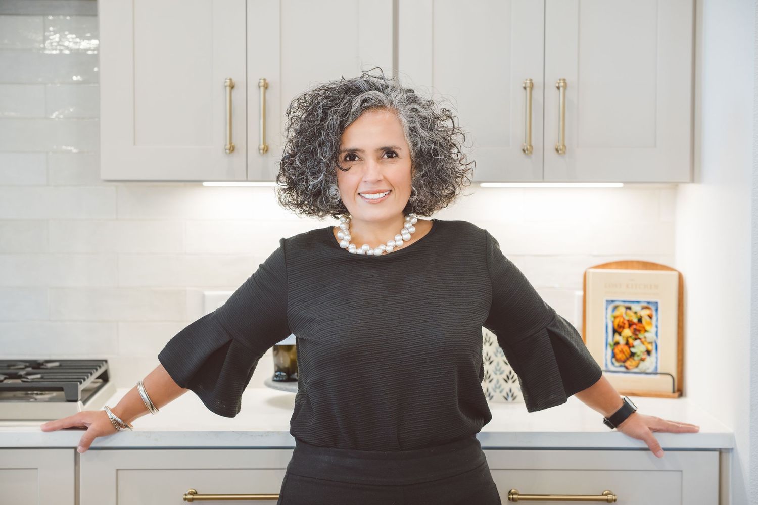 Woman in black smiling while standing in modern white kitchen with marble countertops and gold hardware.