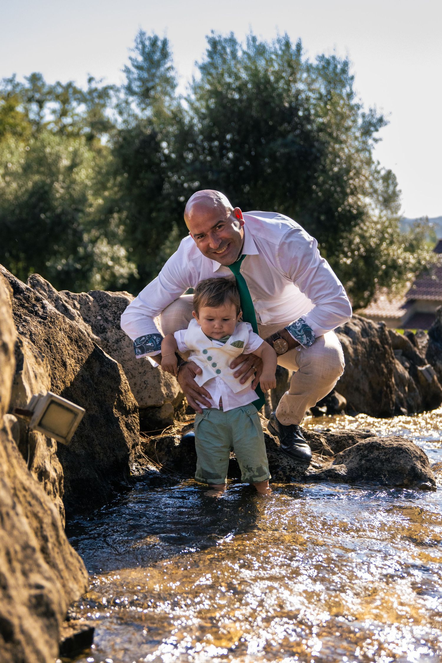 Adult and child explore rocky stream together in white outfits during outdoor photo session.