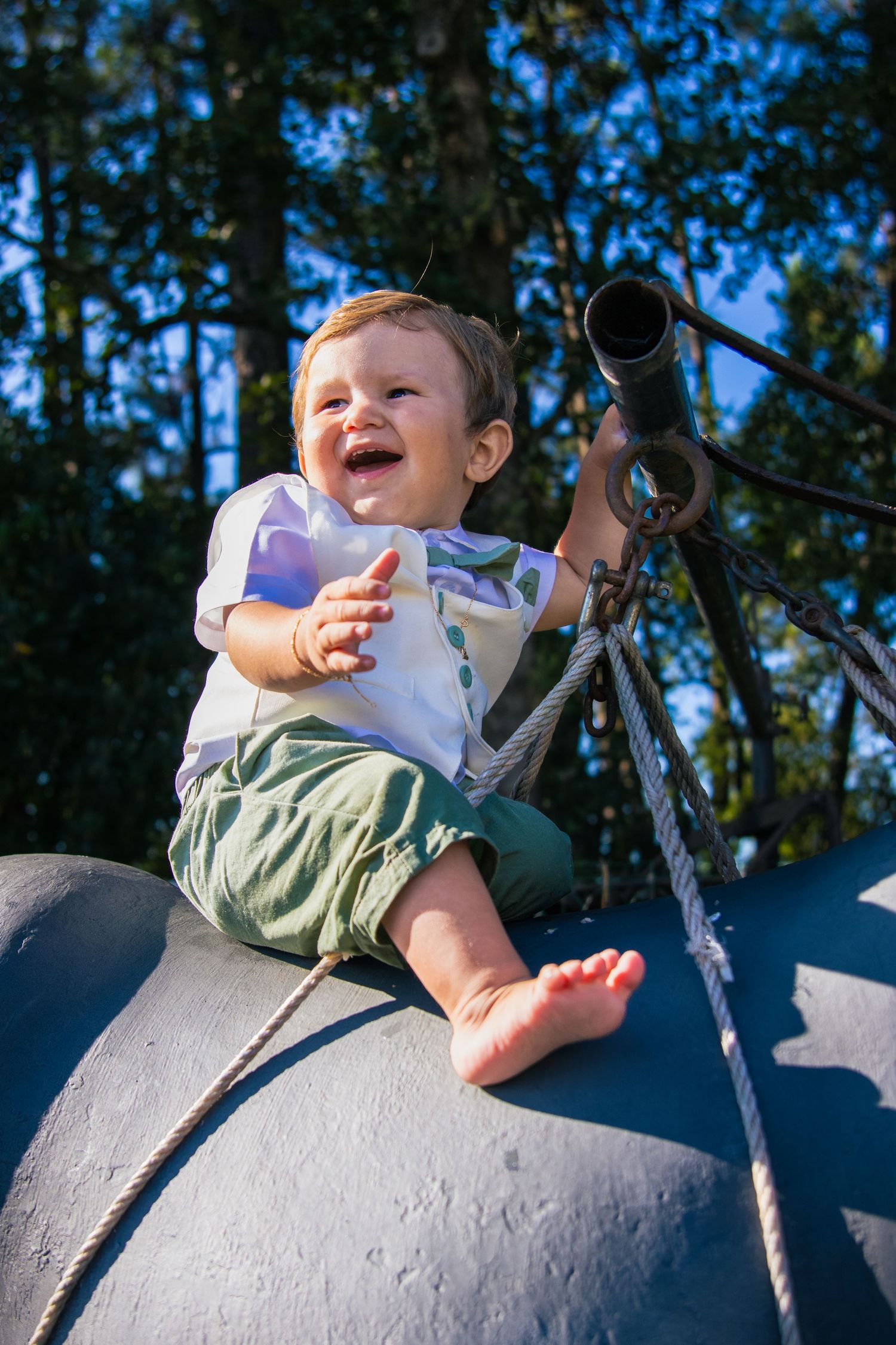 Baby laughing while playing on gray concrete structure outdoors against blue sky and trees.