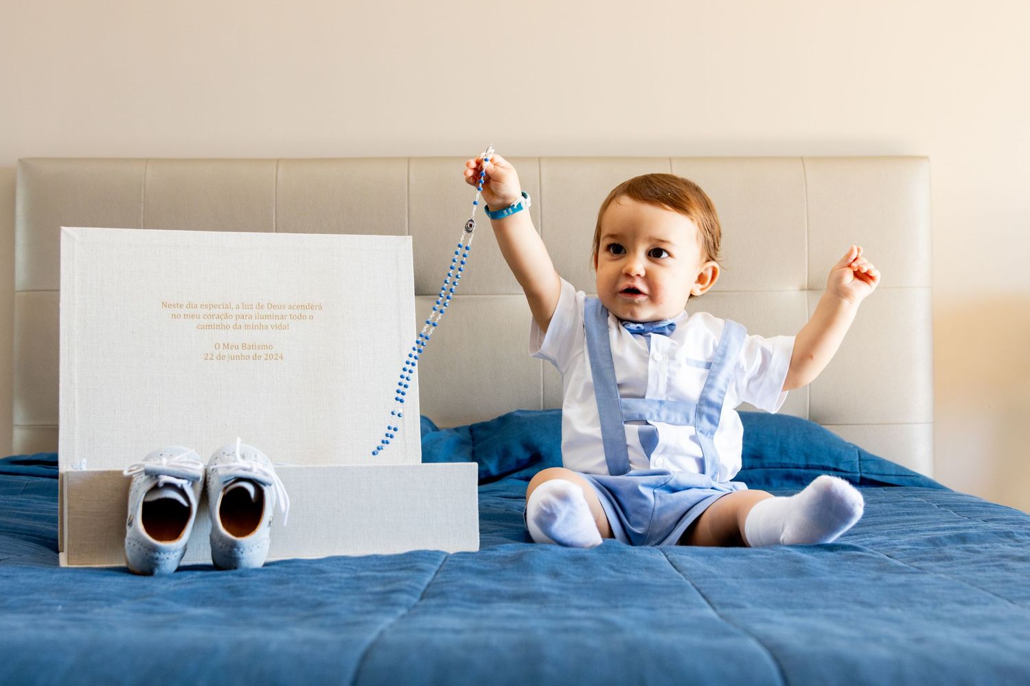 Baby in blue outfit sits excitedly on bed next to small shoes and card.
