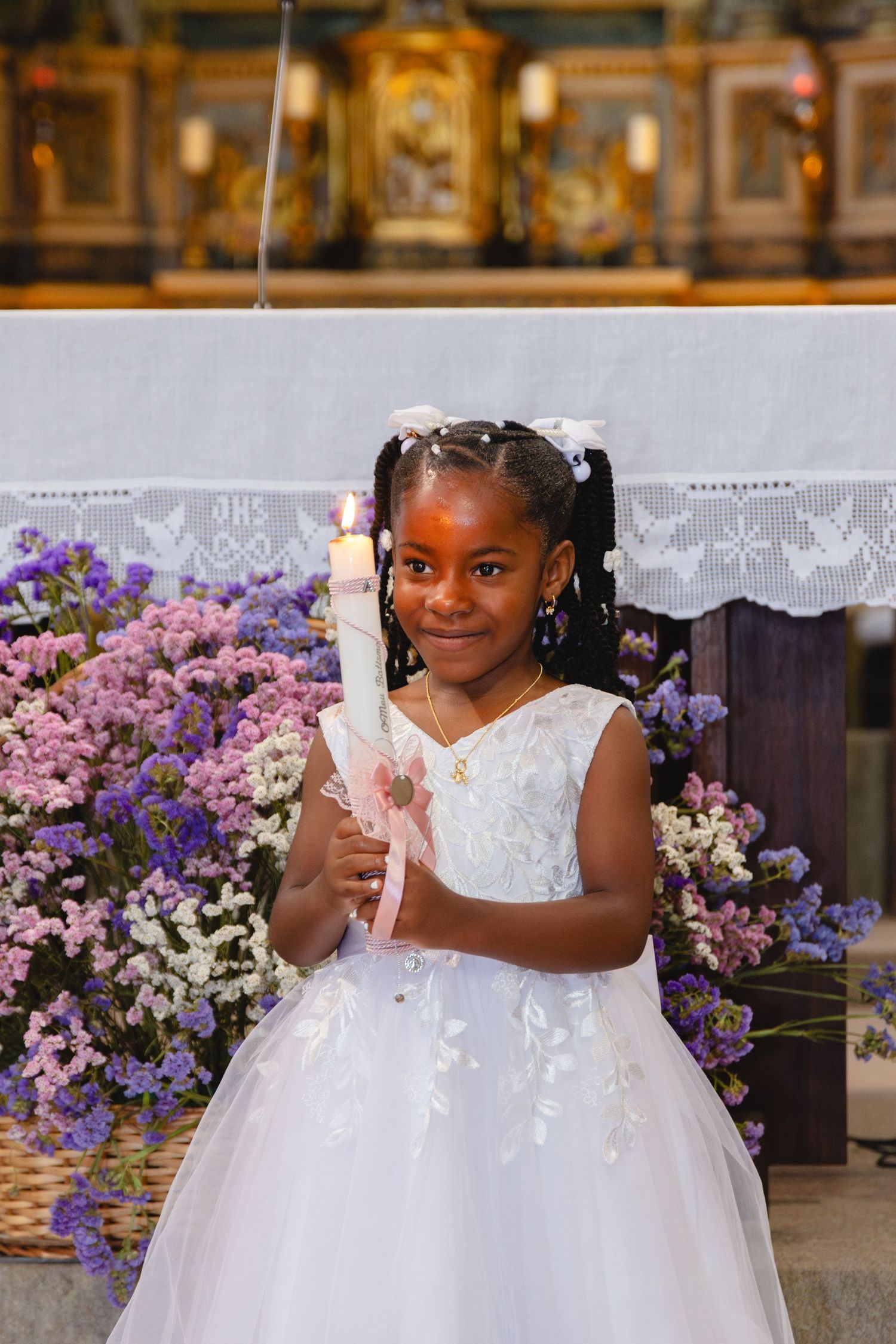 Young flower girl in white dress smiling and posing with purple flowers at elegant wedding ceremony.