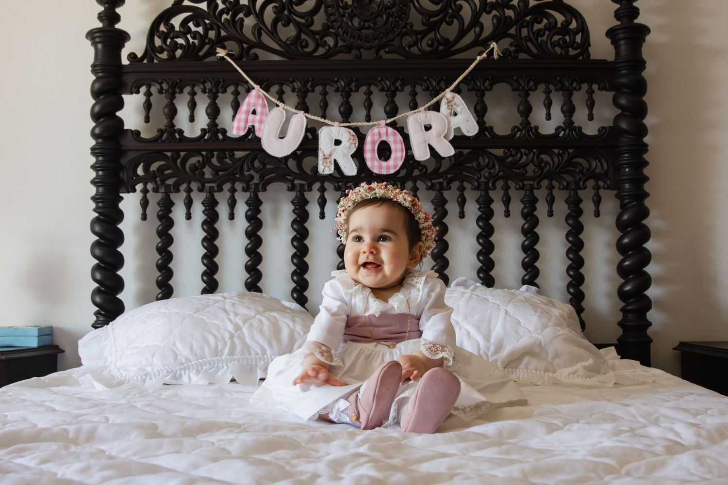 A smiling baby girl sits on a white bed beneath a pink letter banner spelling AURORA on an ornate dark wooden headboard.