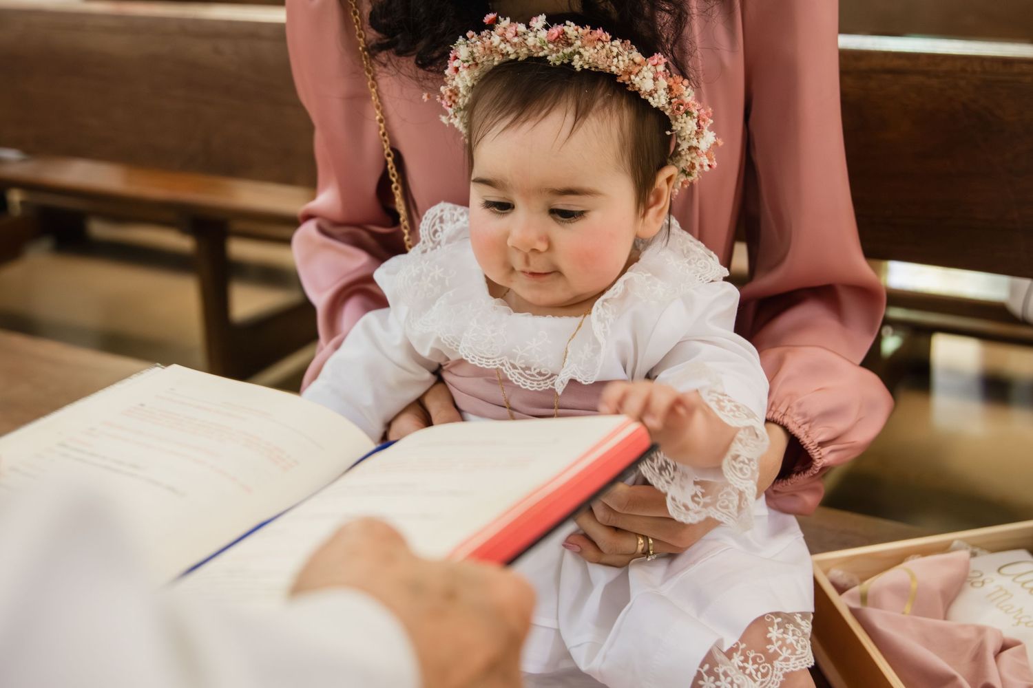 Baby in white lace dress with floral crown touches a book during a christening ceremony in a church.