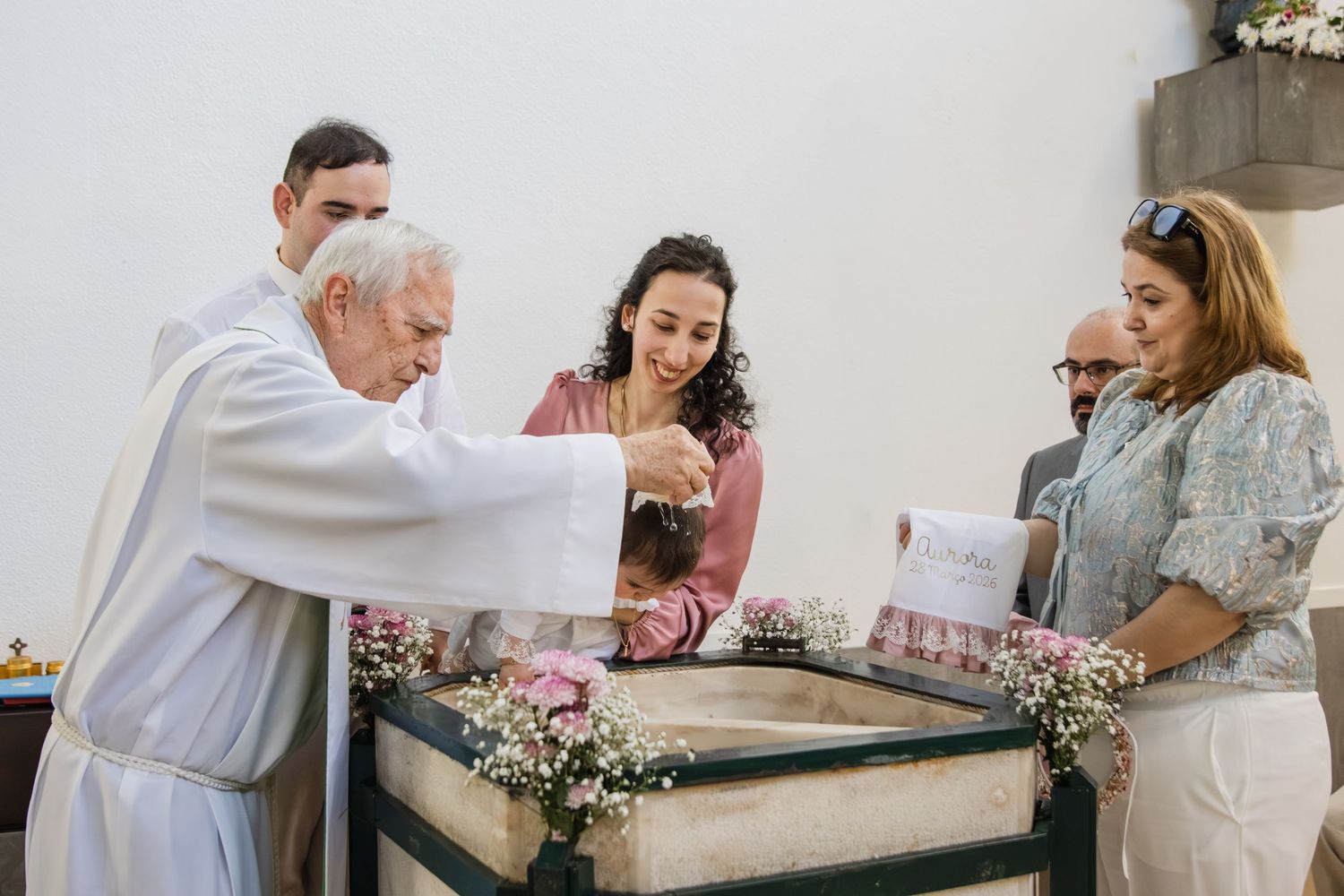 Priest performs Catholic baptism ceremony at font surrounded by family and pink flowers.