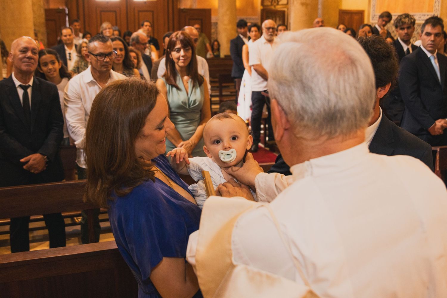 Priest blessing a baby with a pacifier held by a woman in blue dress during a baptism ceremony in a church.