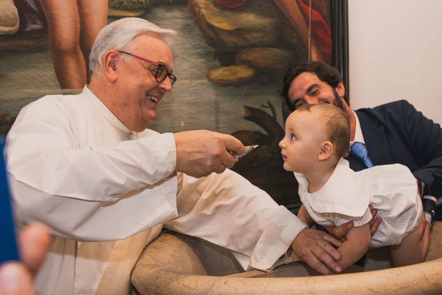 Elderly priest smiles and interacts with a baby held by a man in suit.
