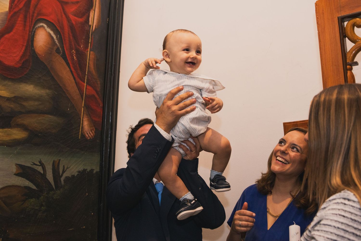 Joyful father lifting smiling baby in white outfit at a baptism, mother watching happily nearby.