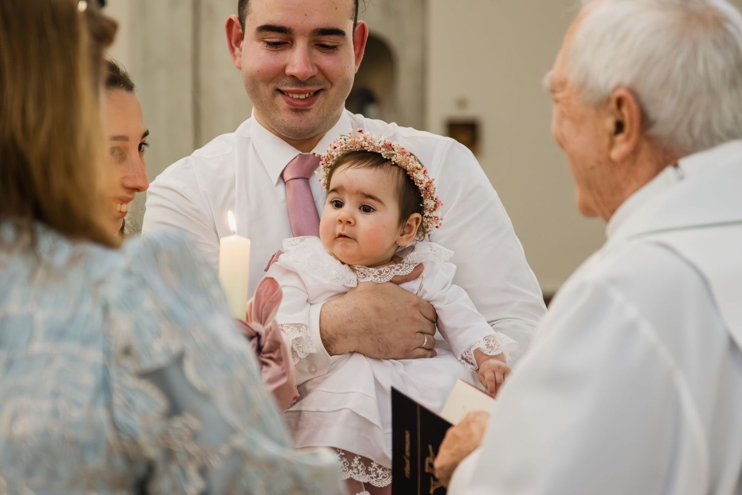 A baby in white christening gown with floral headband held by smiling man during baptism ceremony with priest and candle.