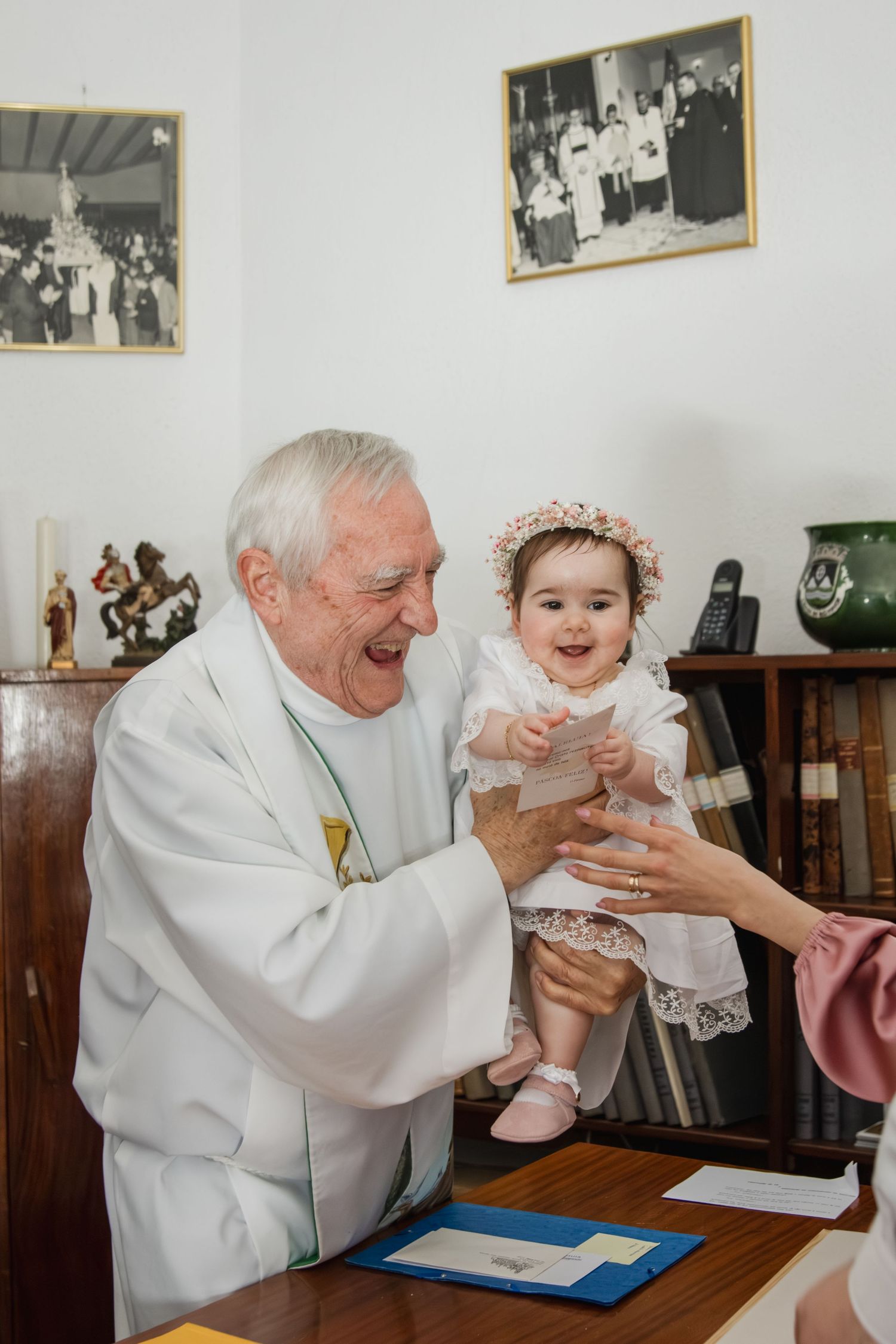 Elderly priest in white robes joyfully holds a smiling baby girl in christening dress and floral bonnet.