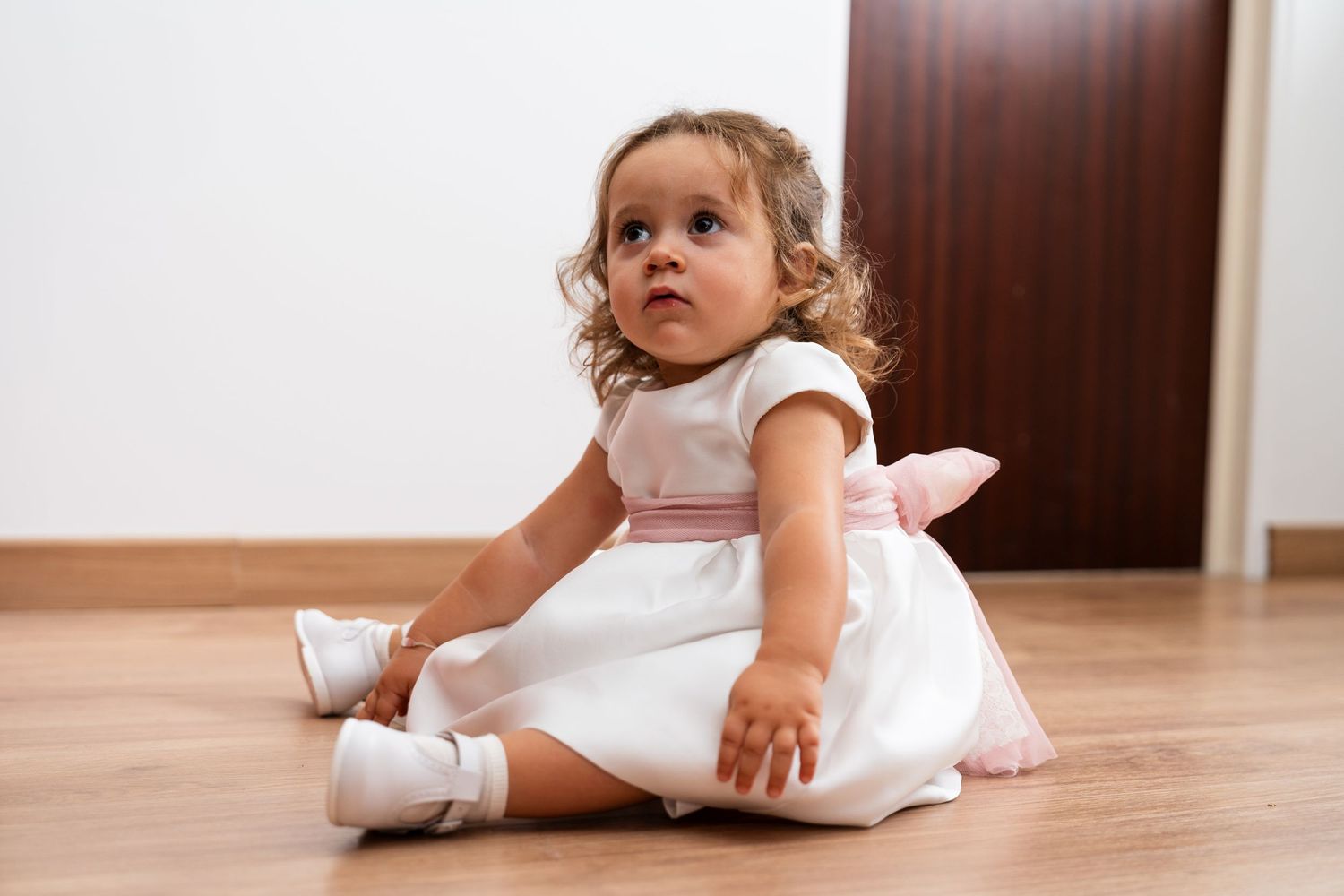 Young attendee in white dress plays on hardwood floor during wedding festivities.