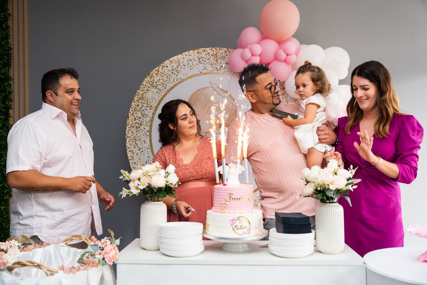 A group gathers around a pink and white birthday cake with candles at an elegant celebration.