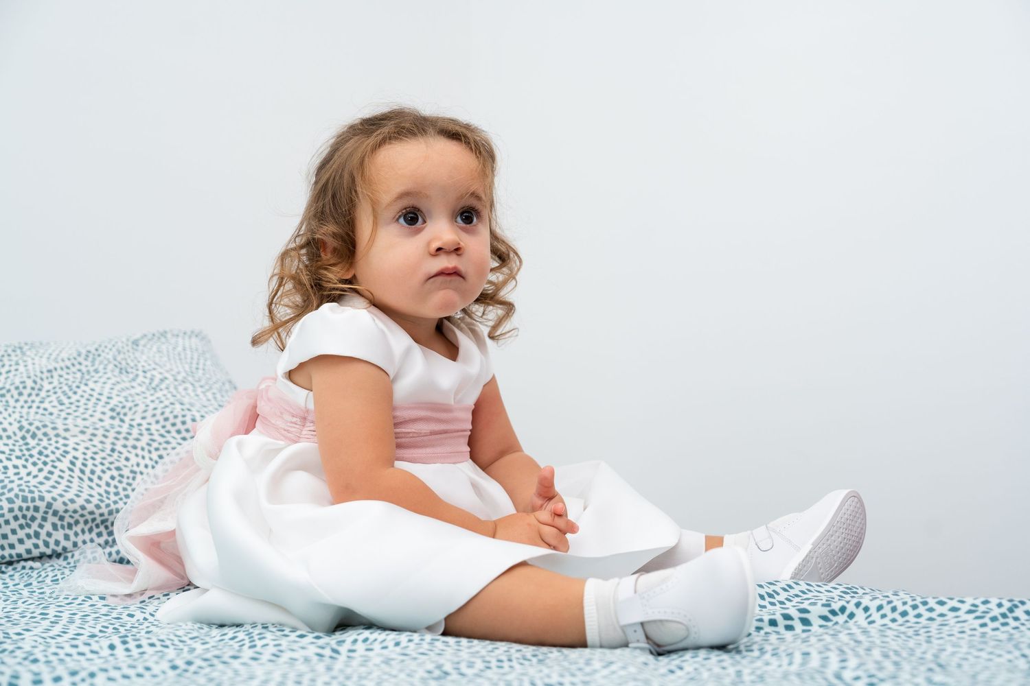 A toddler in a white dress and socks sits on a patterned blue blanket.