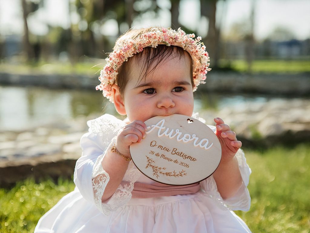 Baby girl in white dress and floral crown chewing a wooden baptism keepsake sign named Aurora outdoors.