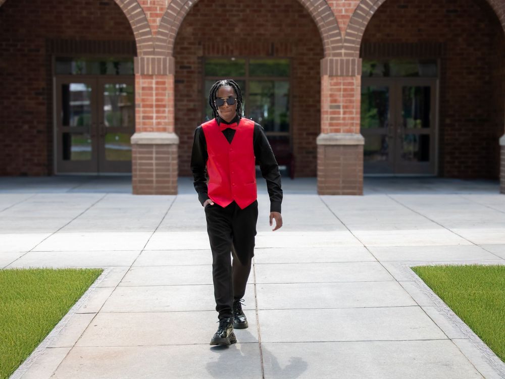 A person wearing a red vest and black suit walks along a concrete path by brick arches and columns.
