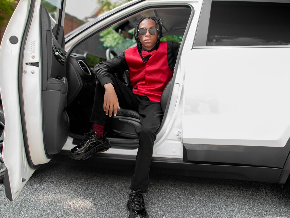 Person wearing red hoodie, sunglasses and black pants sits in driver seat of white car with door open.