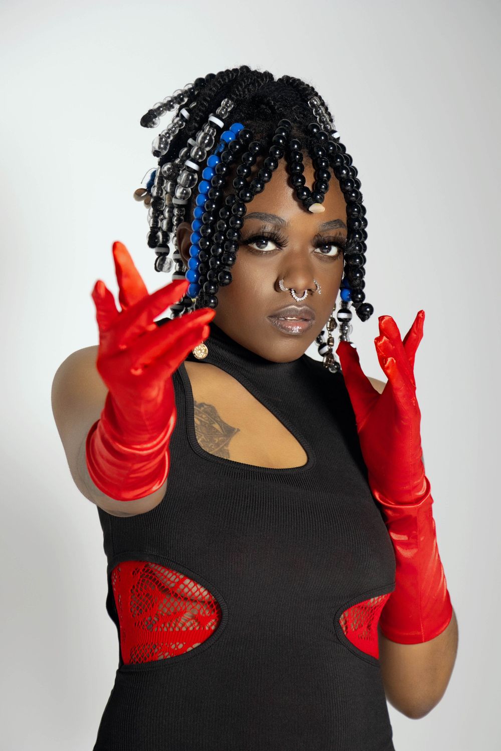A fashion portrait of someone wearing a black cutout dress with red gloves and black braids styled with blue accents.