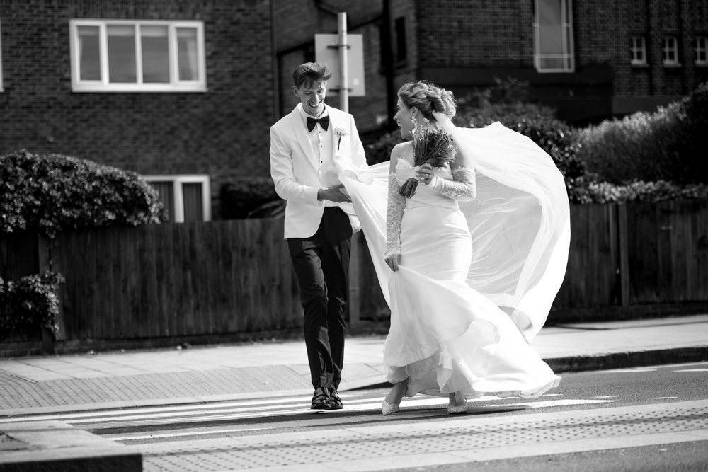 Black and white photo of a couple in wedding attire walking together on a sidewalk with a playful wind blowing the dress.