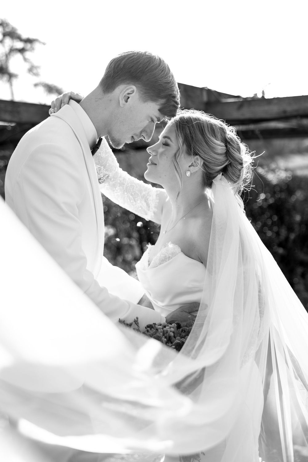 A romantic black and white wedding photograph showing a couple embracing outdoors with the bride's veil flowing in the breeze.