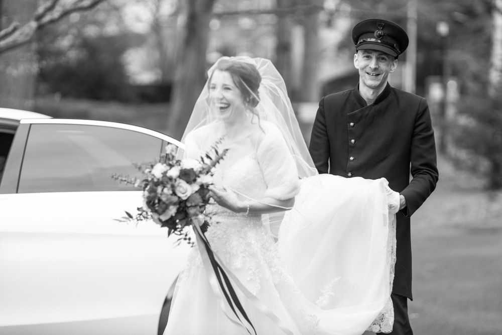 A vintage-style black and white photo of a bride beside a classic car with chauffeur.