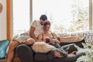 Couple relaxing together with a baby on a comfortable bed in a sunlit bedroom with natural wood window frames.