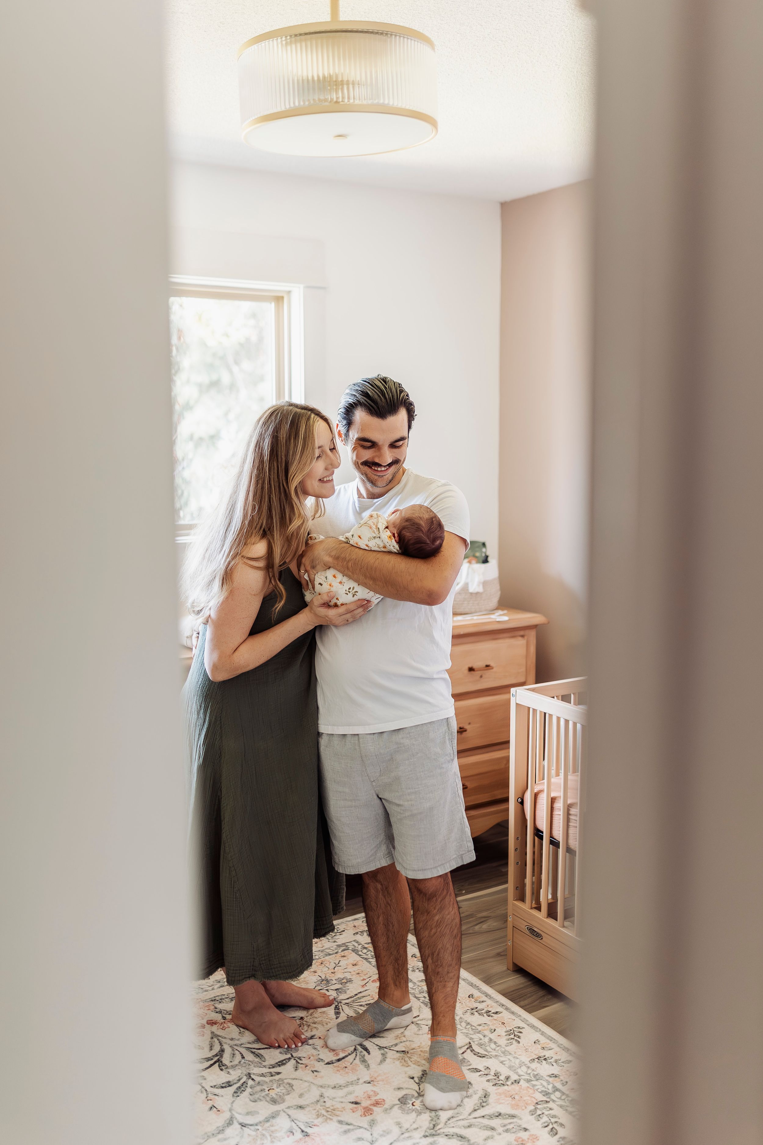 A couple stands together in a bright nursery room with a wooden crib and dresser visible in the background.