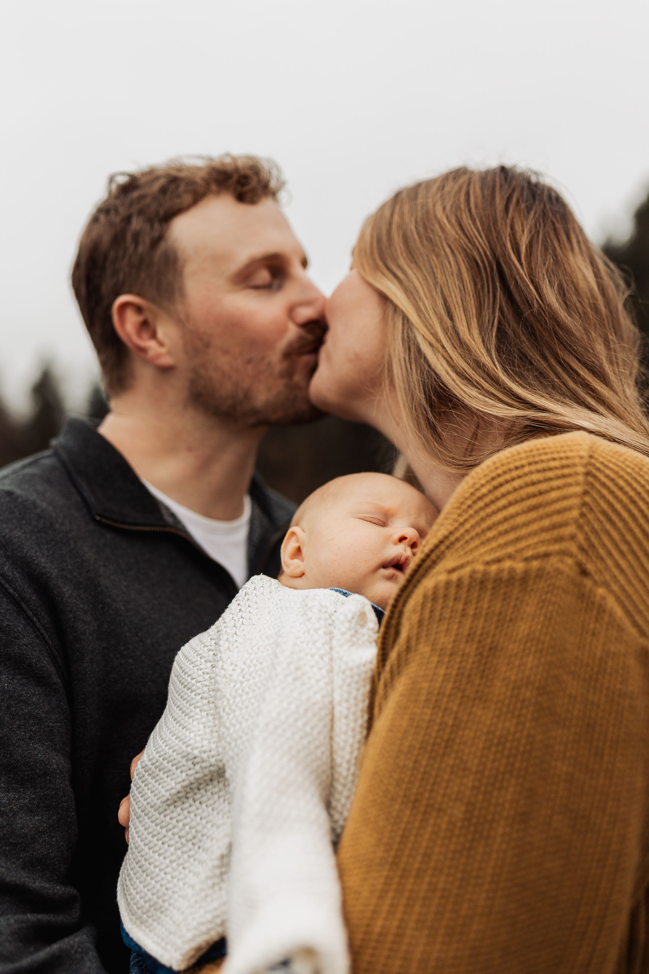 A sleeping baby in white knit clothing is held between two adults in a tender outdoor family moment.