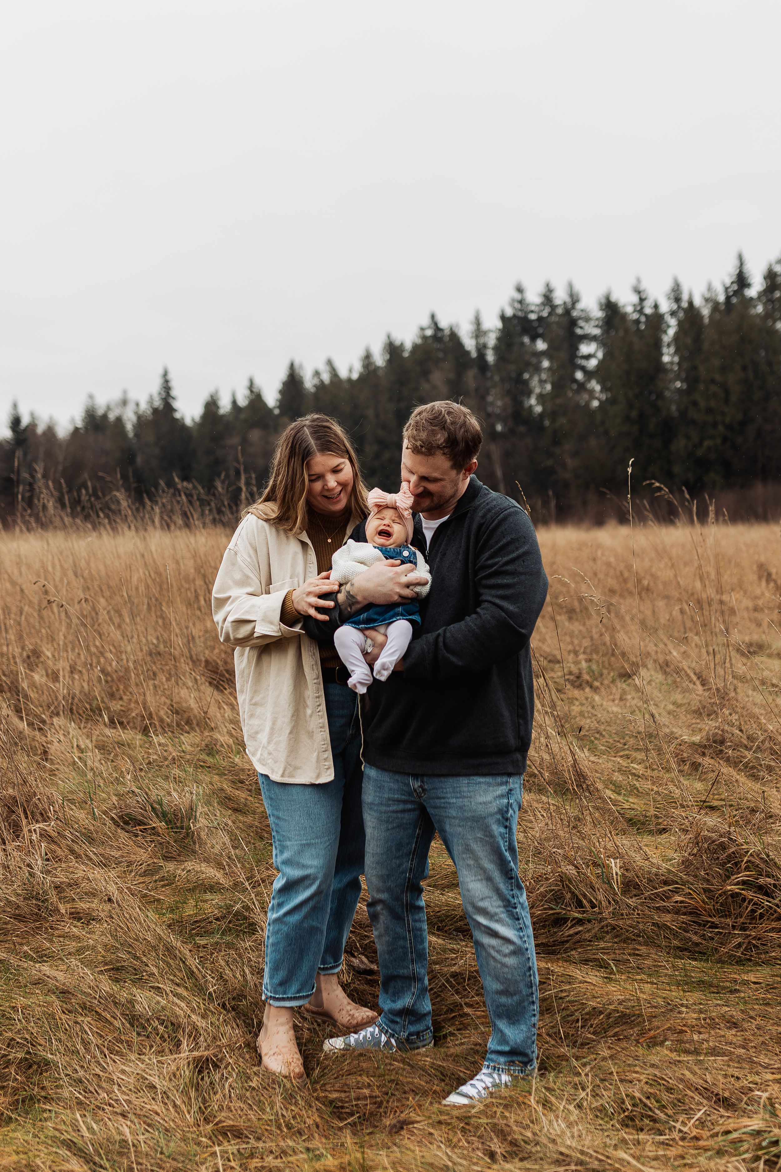 A couple plays with their baby in a golden autumn field surrounded by evergreen trees in the distance.