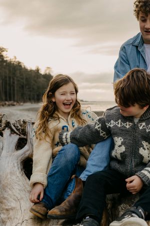 Children laughing while sitting on driftwood at a beach with forest backdrop.