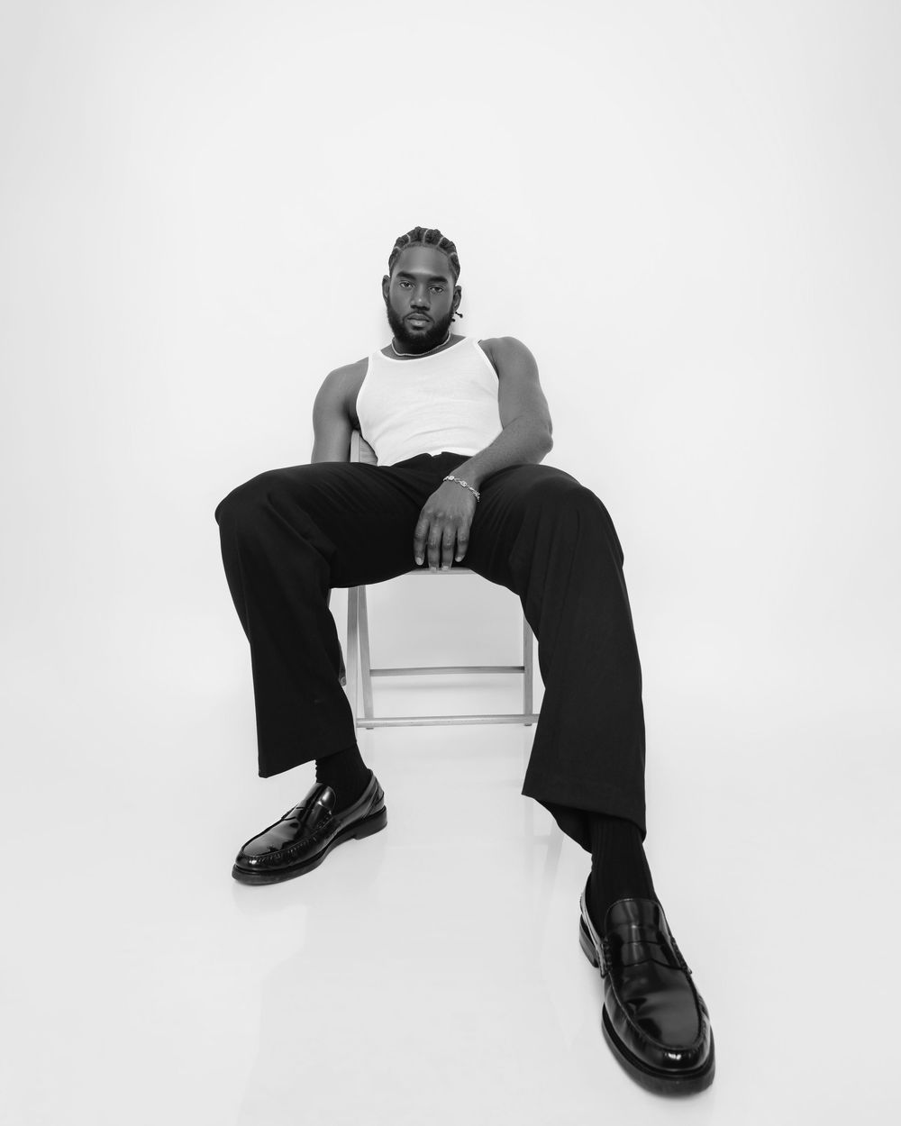 Black and white photo of a man in a white tank top and black pants sitting confidently on a stool.