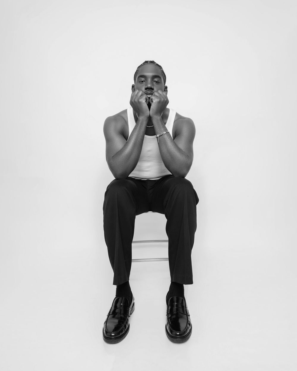 Black and white portrait of a muscular man sitting on a stool with hands raised to his chin.