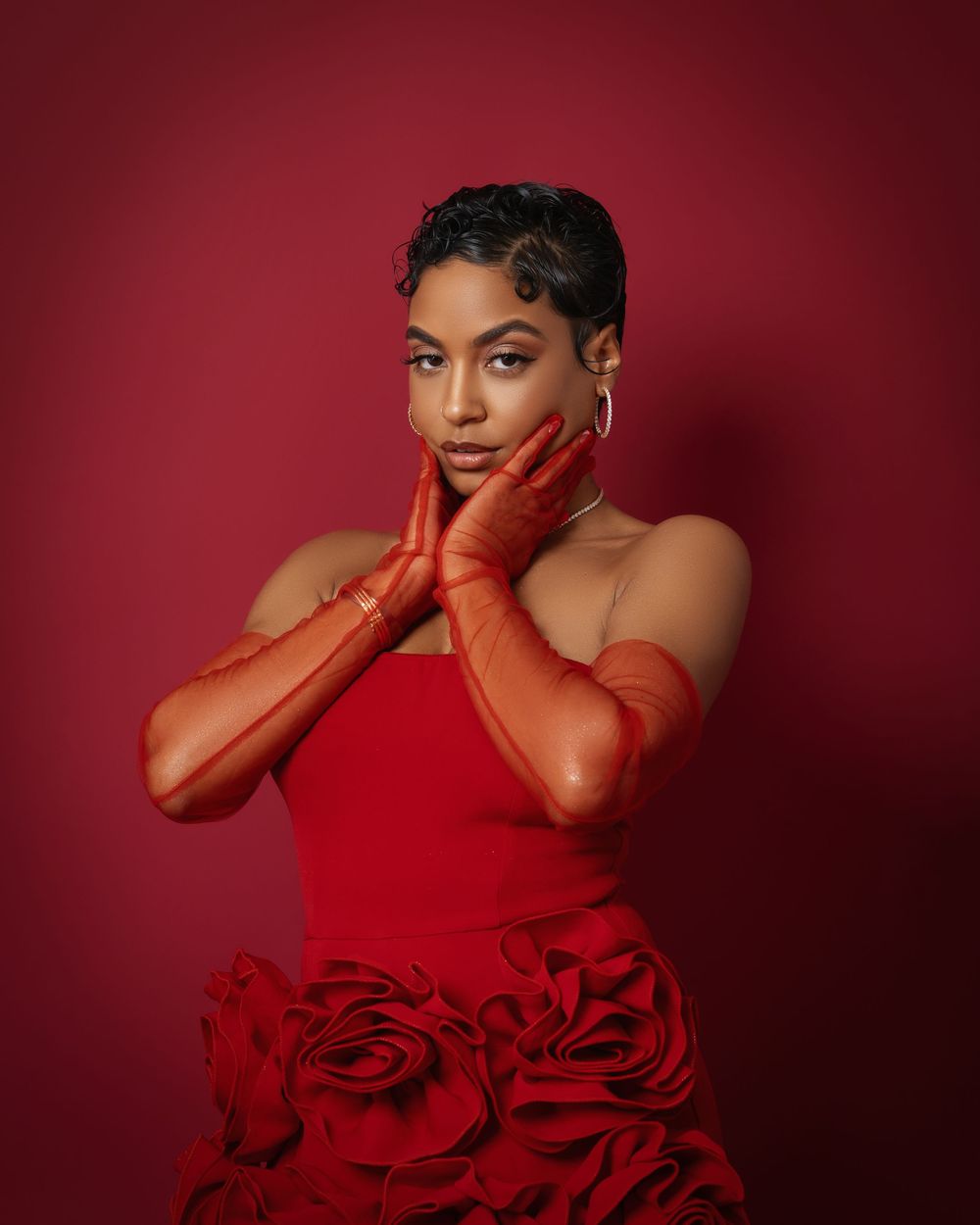 A woman in a red strapless floral dress and long red gloves poses against a deep red background.