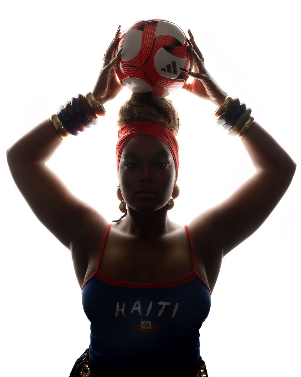 Silhouetted woman in Haiti jersey balances Adidas soccer ball overhead on white background.