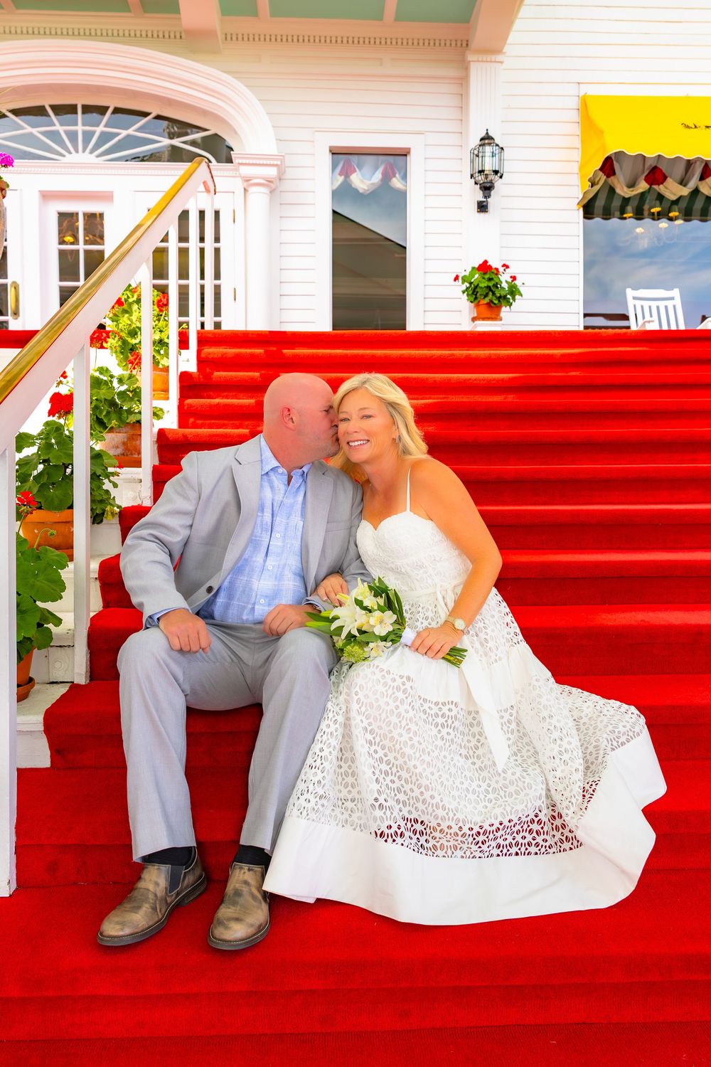A couple in wedding attire sits together on bright red carpeted stairs outside a white building.