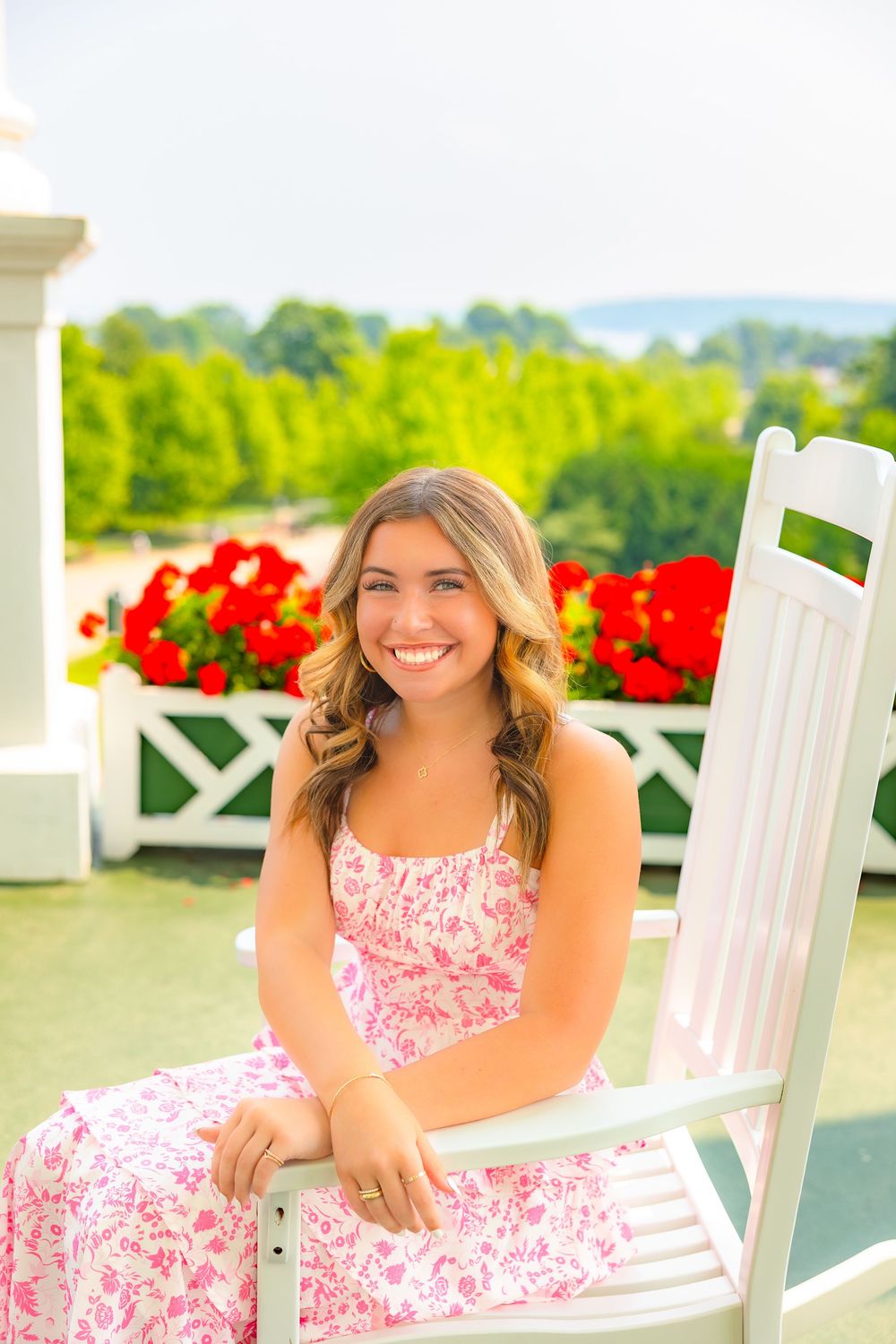 Person in pink and white floral dress relaxes on white rocking chair with red flowers in background on sunny porch.