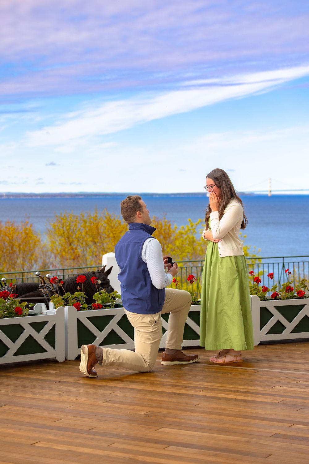 A romantic proposal moment on a waterfront deck with ocean views and colorful flowers in the background.