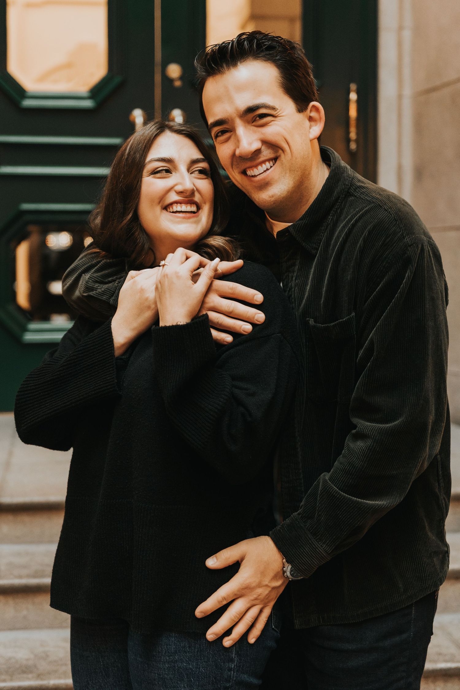 A joyful couple wearing black outfits laughs together while embracing outdoors on a city street.