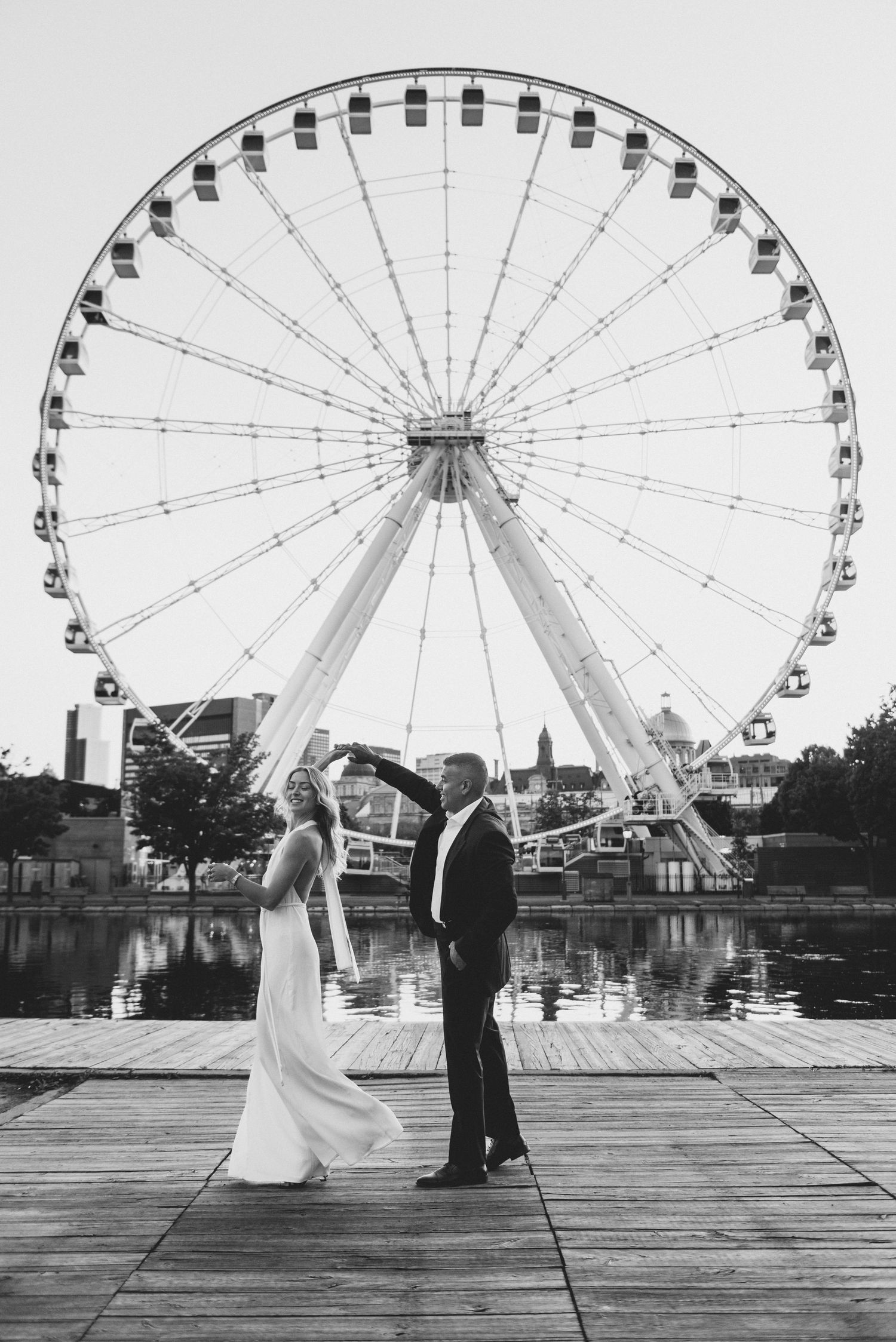 A couple stands in silhouette on a wooden pier with a dramatic ferris wheel in the background at dusk.
