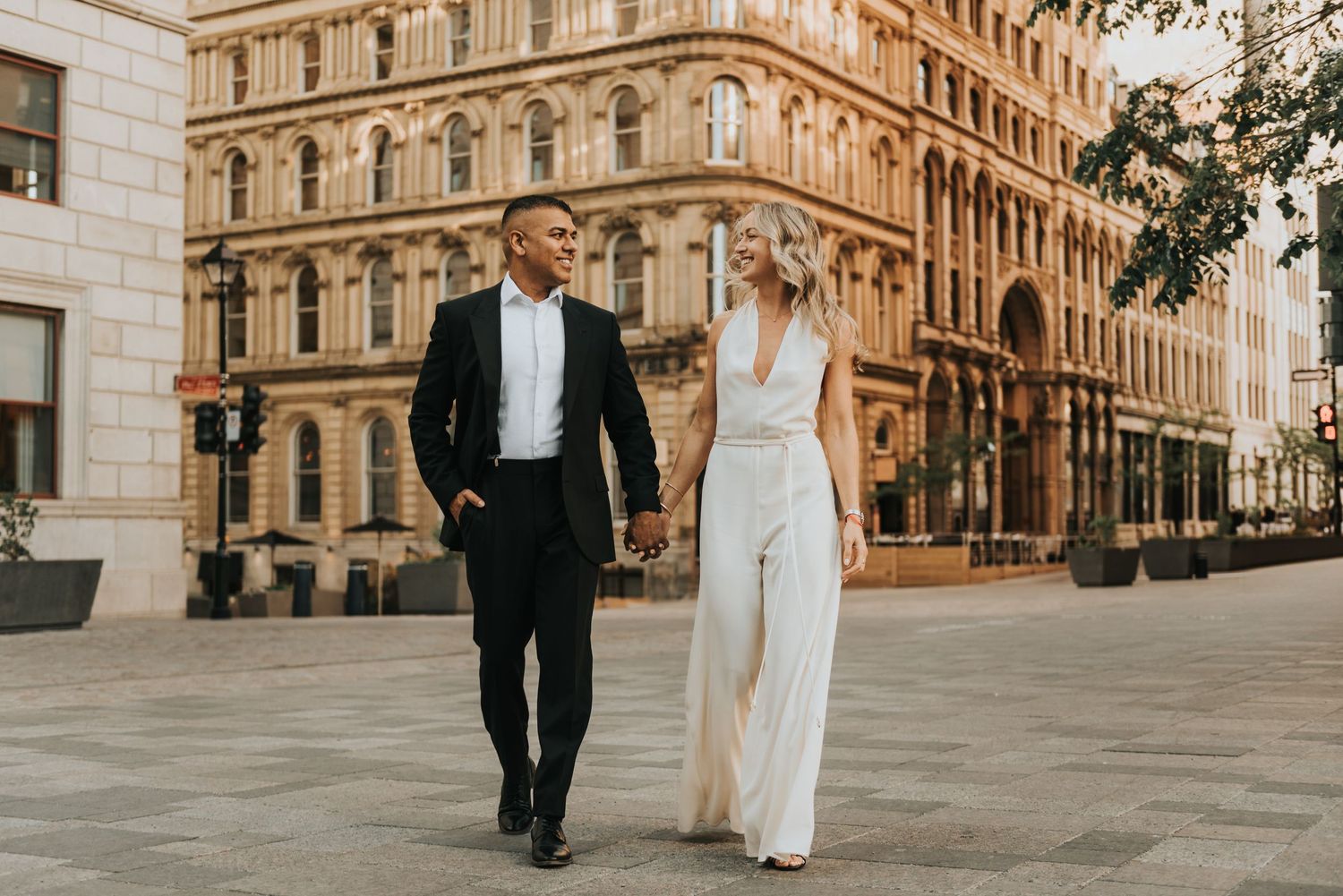A couple in formal attire walks hand in hand along a city street with grand historic architecture in the background.