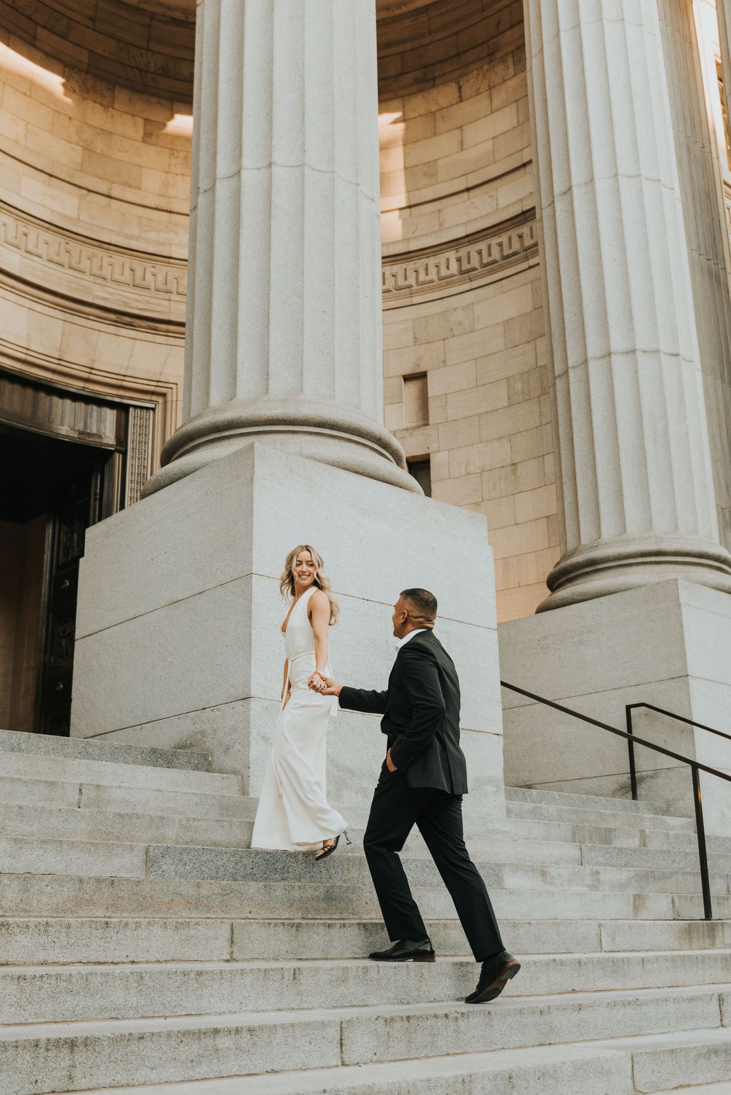 Elegant bride and groom in formal attire dancing on grand stone steps with classic white columns in background.