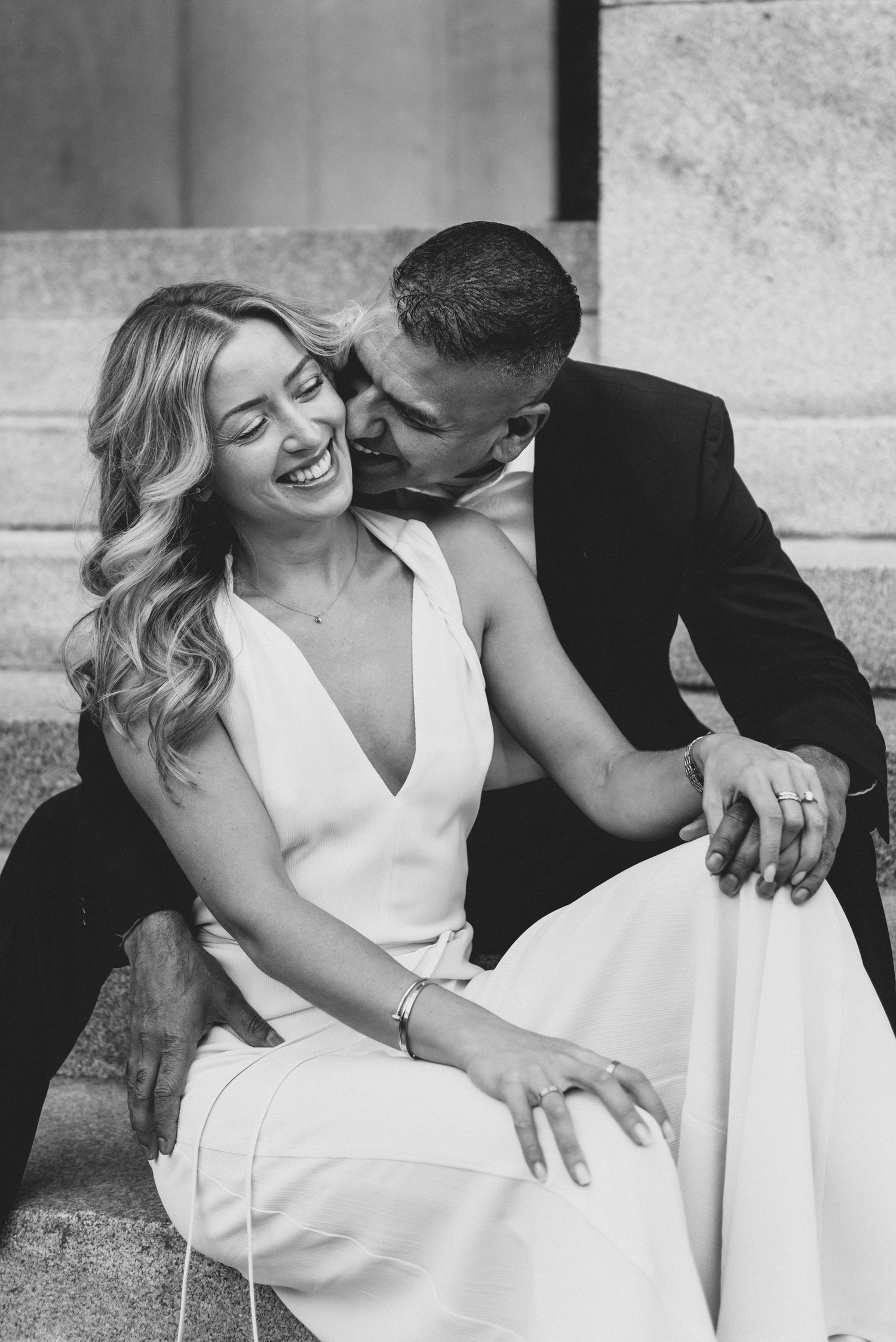 A loving couple shares an intimate black and white moment on stone steps, with the man kissing his partner's cheek.