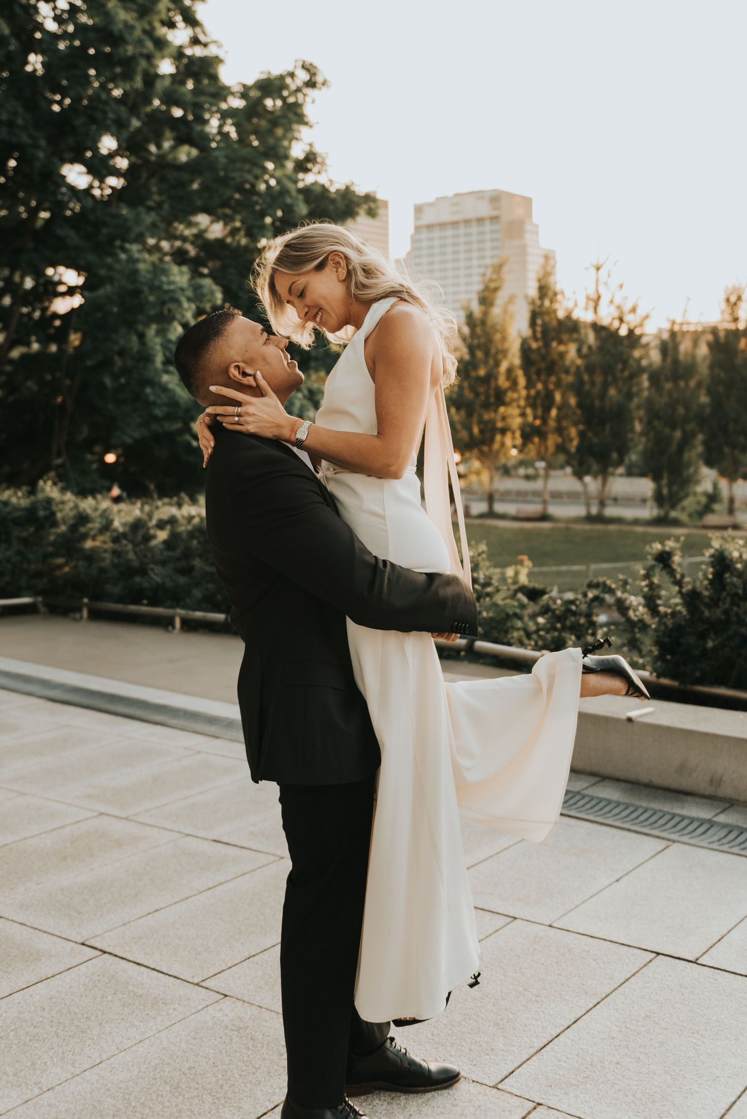 A romantic newlywed couple embraces affectionately at sunset in an urban garden setting with city buildings in background.