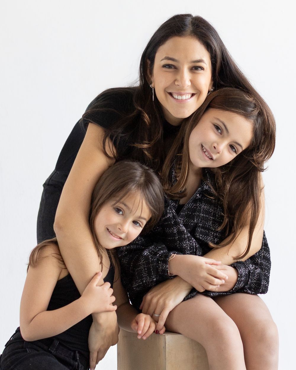 A smiling woman in black poses with two young girls in a bright studio portrait setting.
